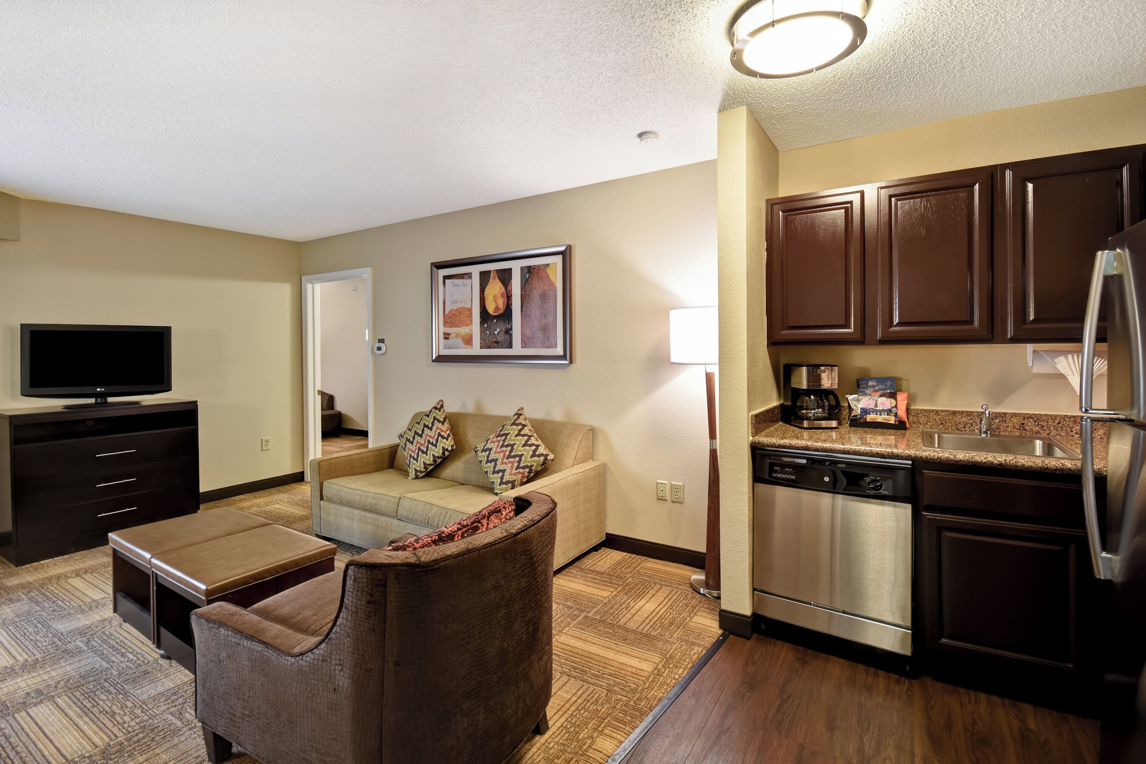 A modern living room with a flat-screen TV, a sofa, a coffee table, and a kitchen area with a dishwasher and refrigerator.