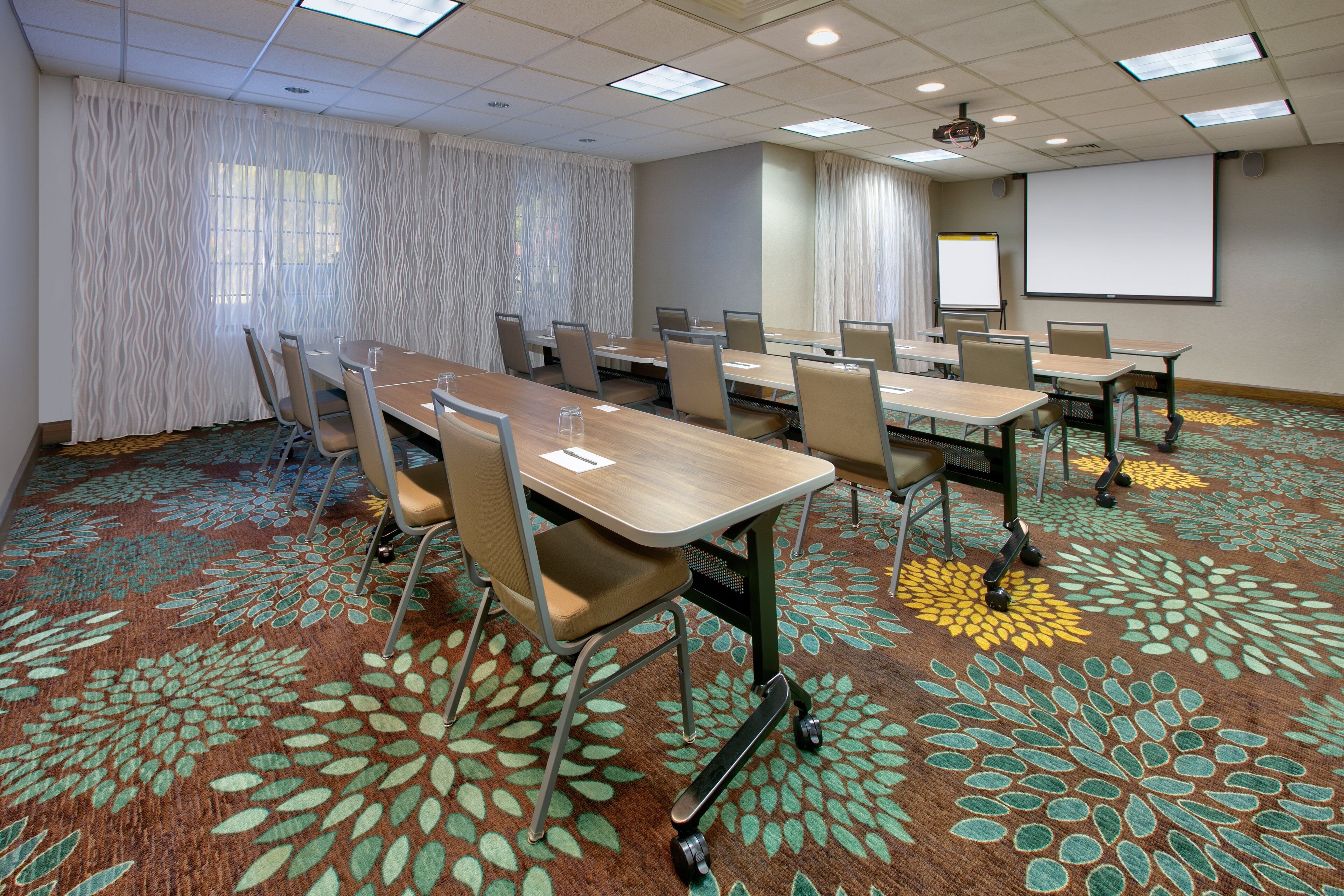 A conference room with a long table, chairs, a projector screen, and a carpet with a floral pattern.