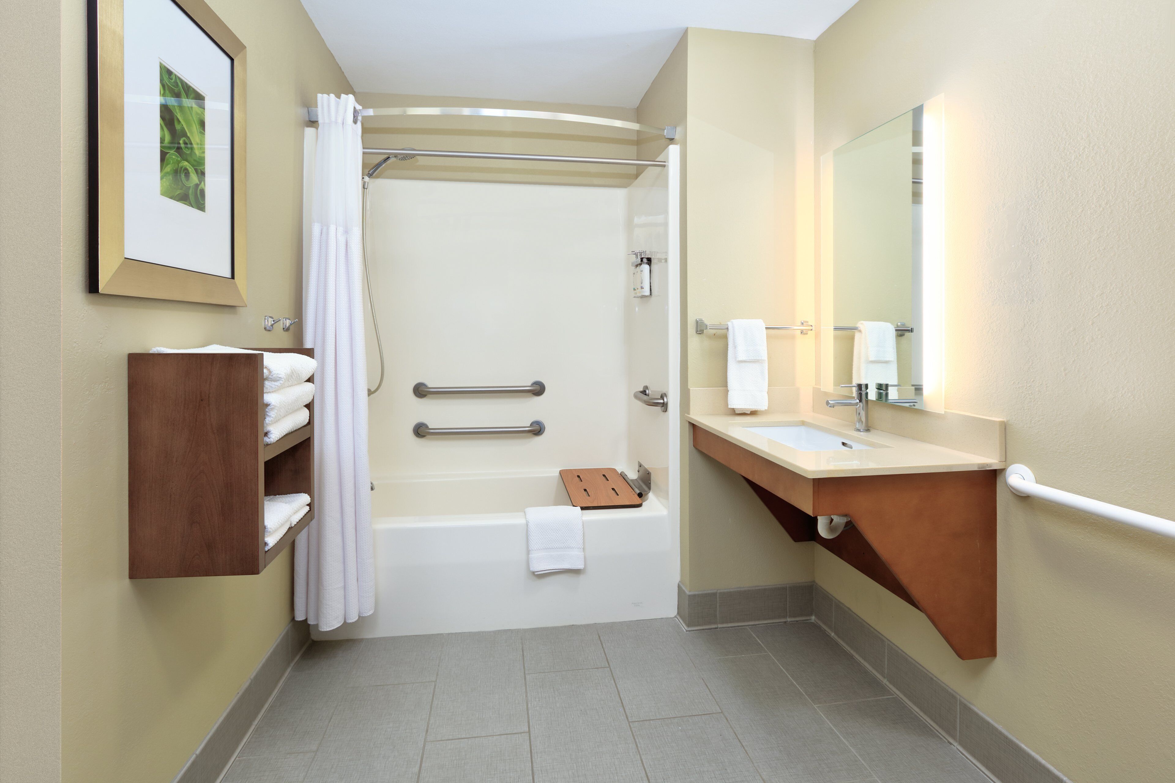 A modern bathroom with a bathtub, shower, and sink. There are handrails, a towel rack, and a wooden shelf with towels.