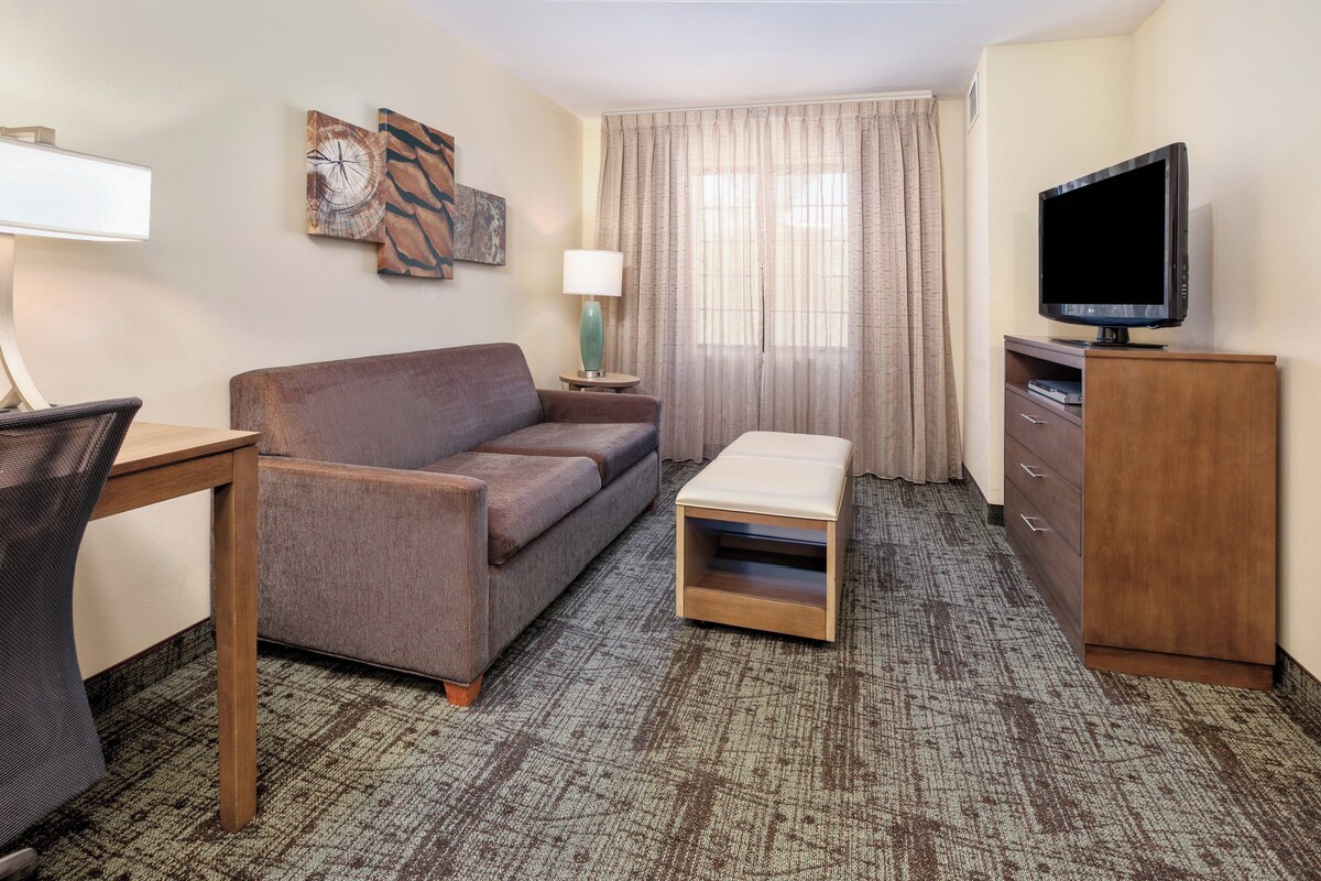 A hotel room with a sofa, a small table, a TV on a cabinet, a lamp, and a window with curtains.