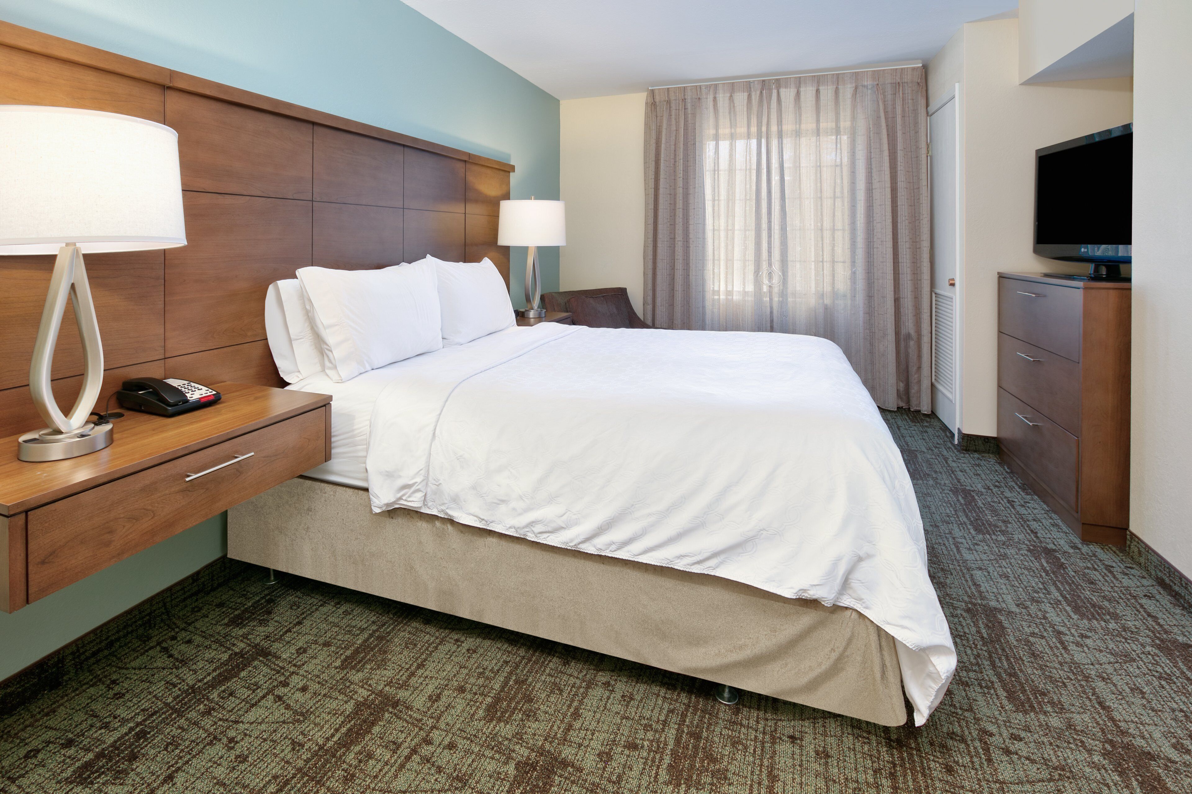 A hotel room with a large bed, a wooden headboard, a desk with a lamp, a TV, and a wardrobe.