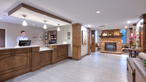 A reception area with a wooden reception desk, a stone fireplace, and pendant lighting. - Staybridge Suites Corning by IHG (Corning)