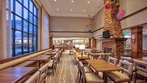 A spacious hotel dining area with long wooden tables and cushioned chairs, large windows, and a stone fireplace. - Staybridge Suites Corning by IHG (Corning)