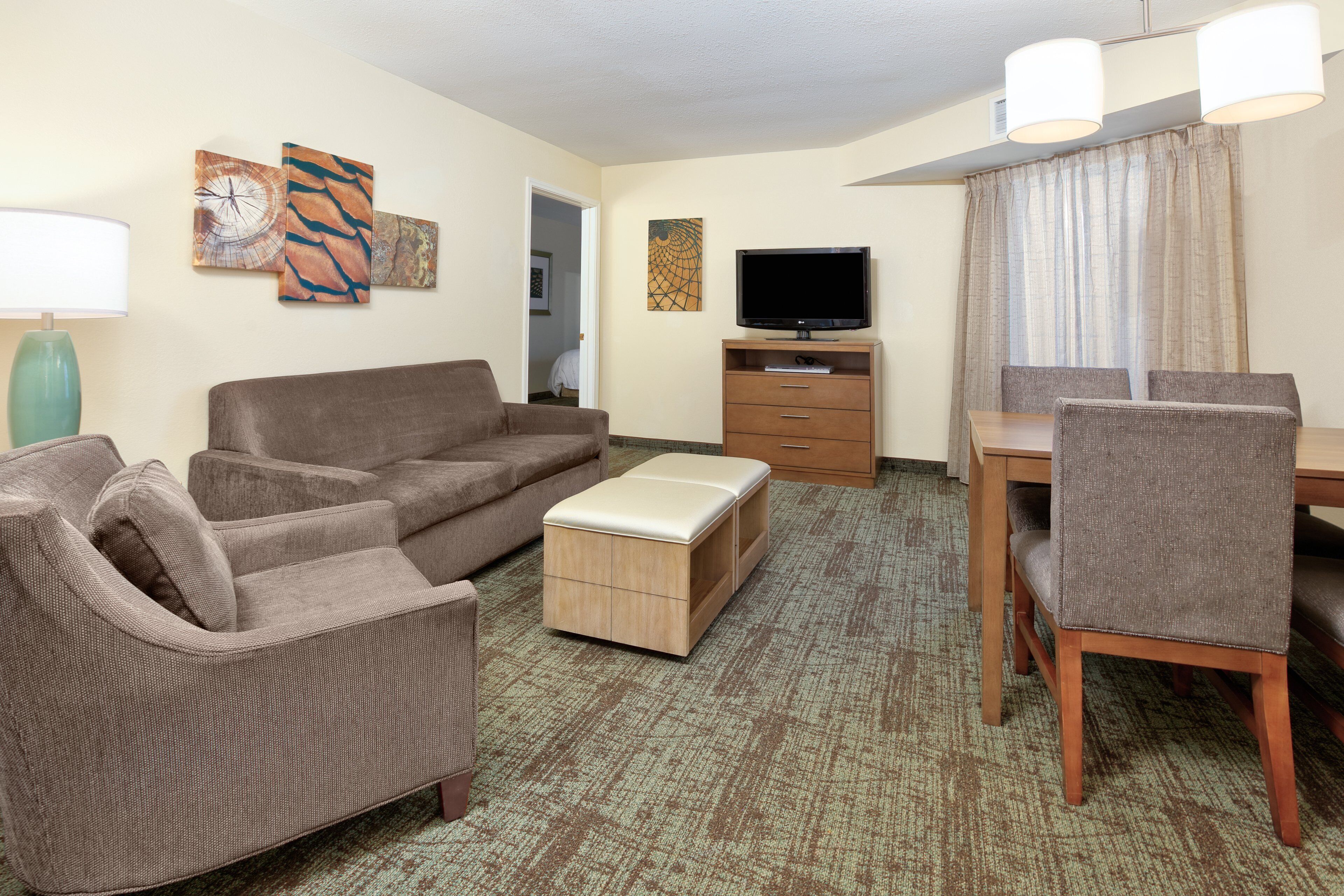 A hotel room with a brown sofa, a wooden ottoman, a TV on a stand, a dining table with chairs, and artwork on the walls.
