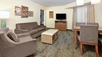 A hotel room with a brown sofa, a wooden ottoman, a TV on a stand, a dining table with chairs, and artwork on the walls.