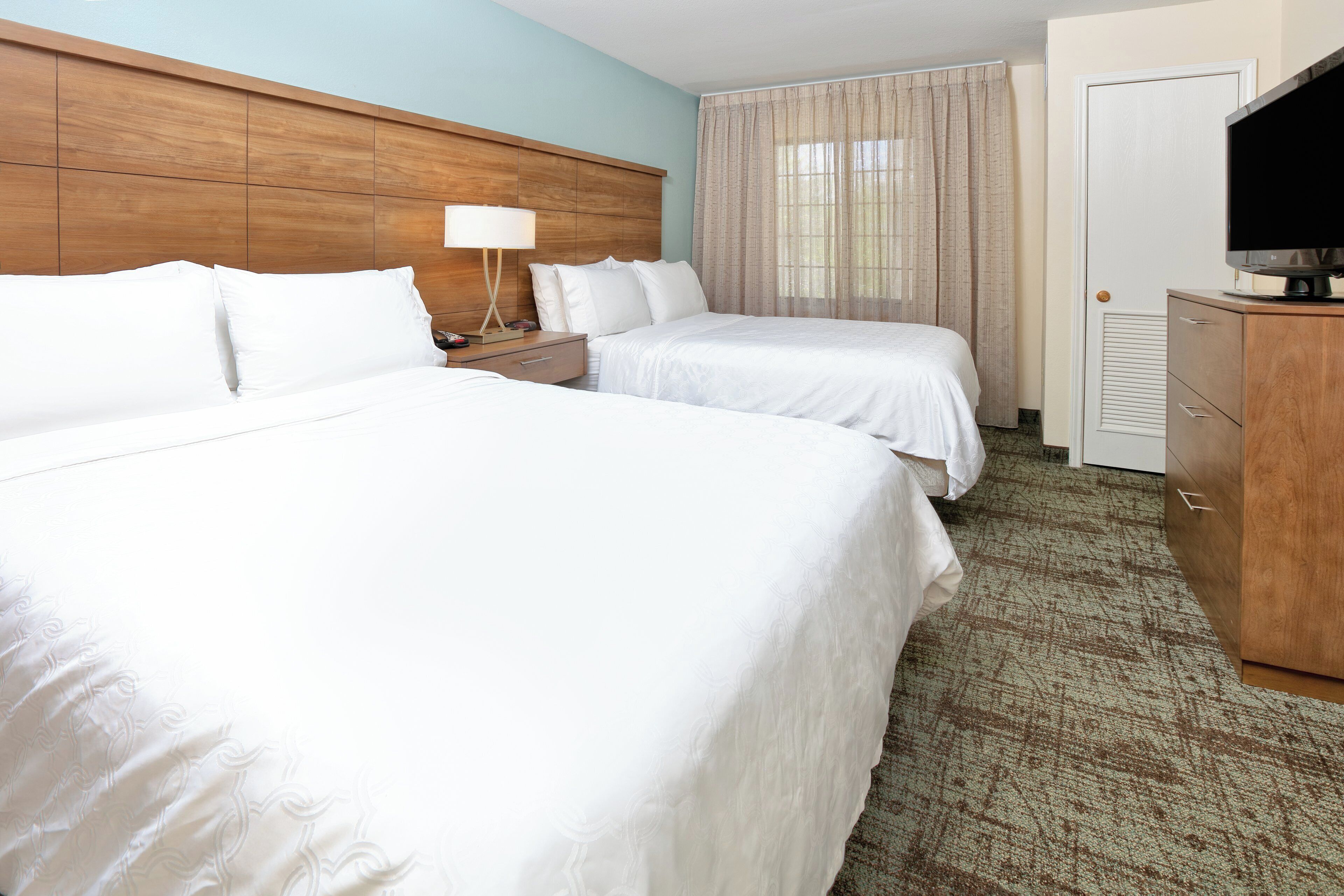 A hotel room with two beds, a nightstand, a lamp, a television, and a wooden headboard.