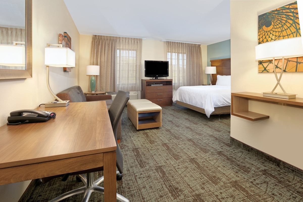 A hotel room with a desk, chair, bed, TV, and a painting on the wall.