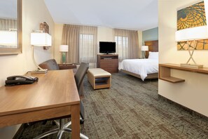 A hotel room with a desk, chair, bed, TV, and a painting on the wall.