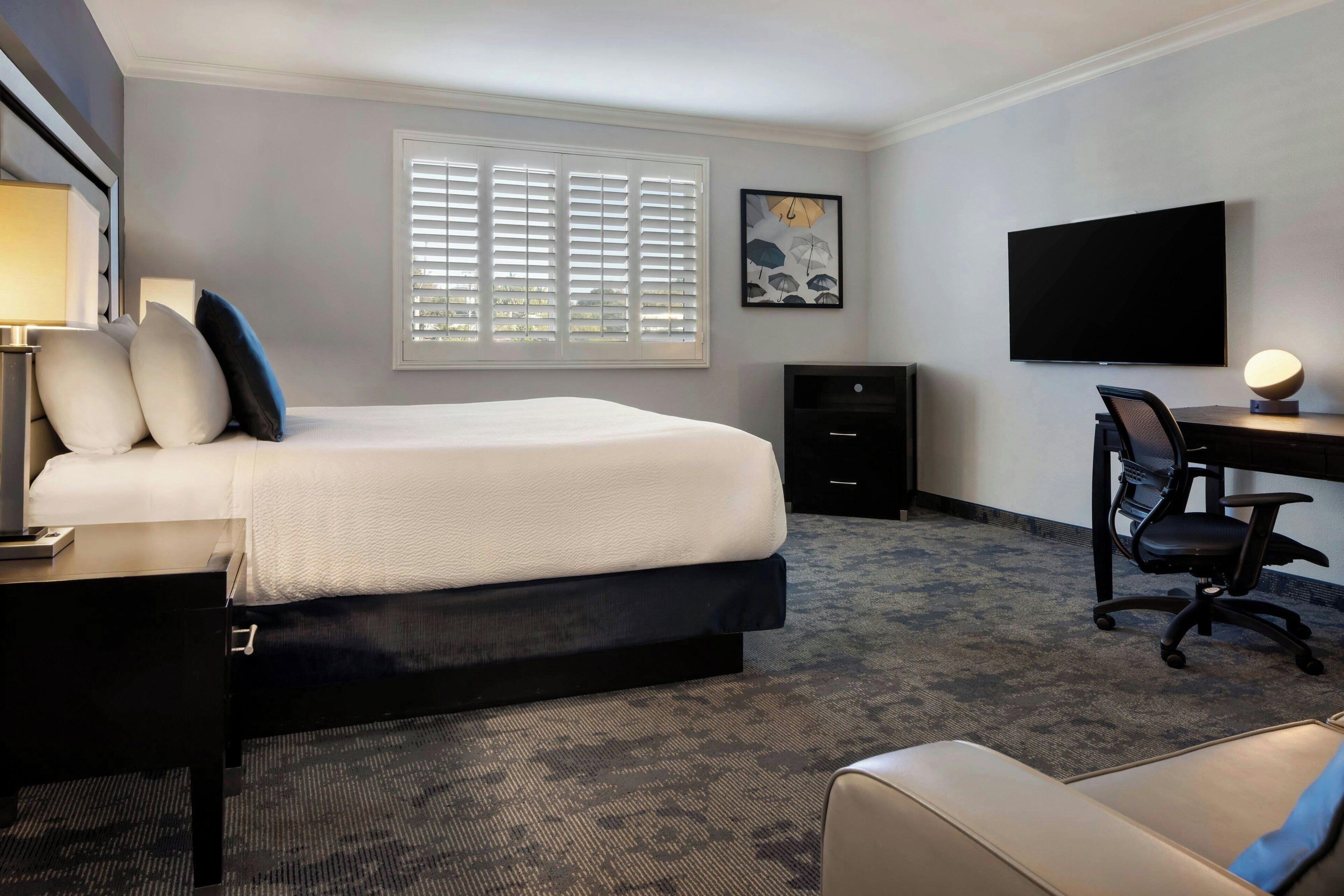 A hotel room with a bed, desk, chair, TV, and a window with blinds.