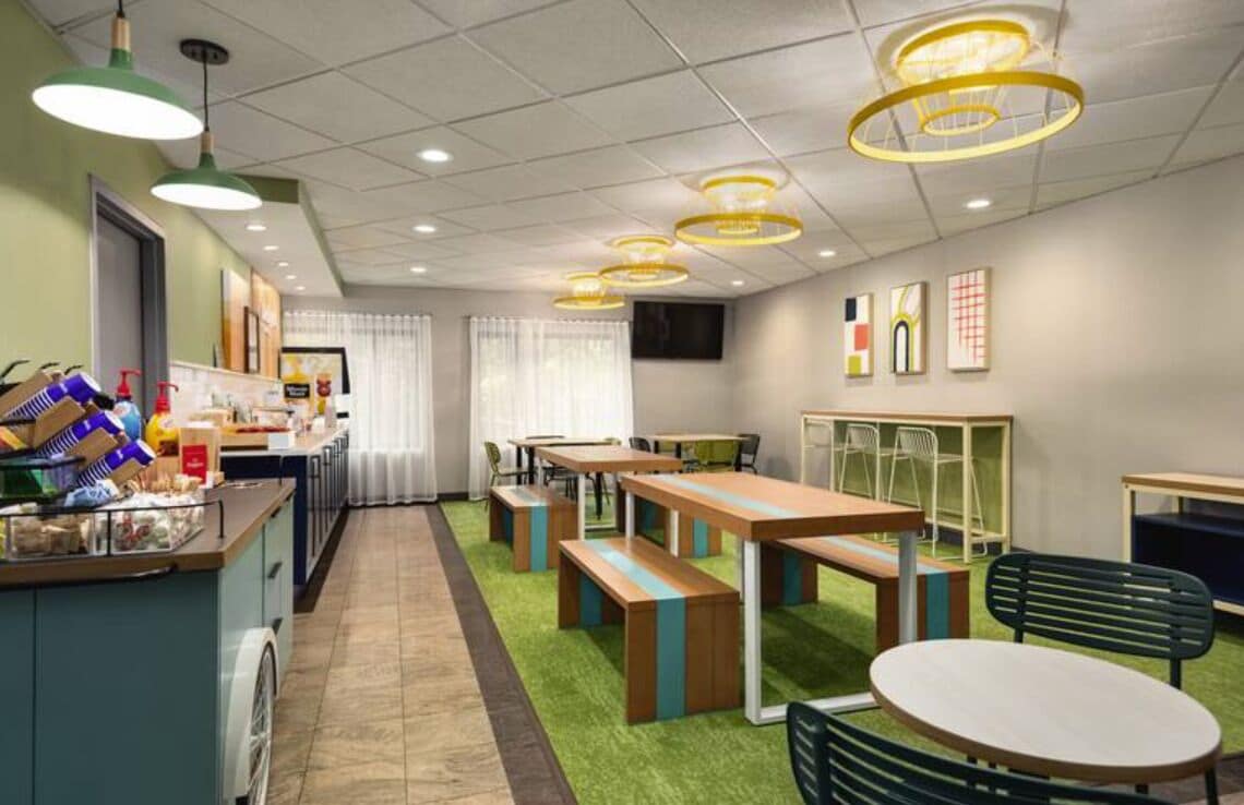 A modern dining area with green artificial grass, wooden tables and benches, and pendant lighting.