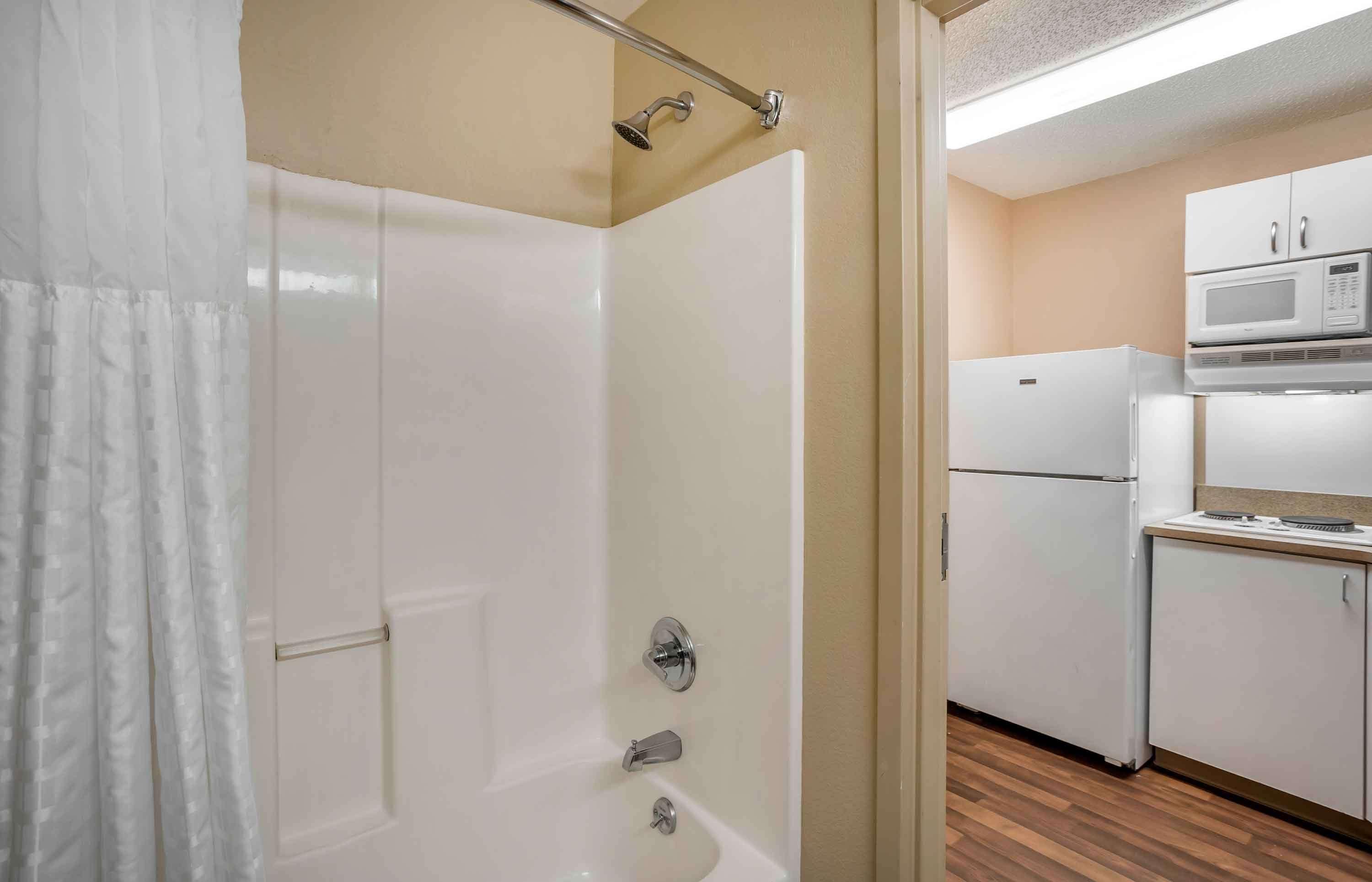 A bathroom with a shower, toilet, and sink. There is a kitchen area with a refrigerator and microwave.