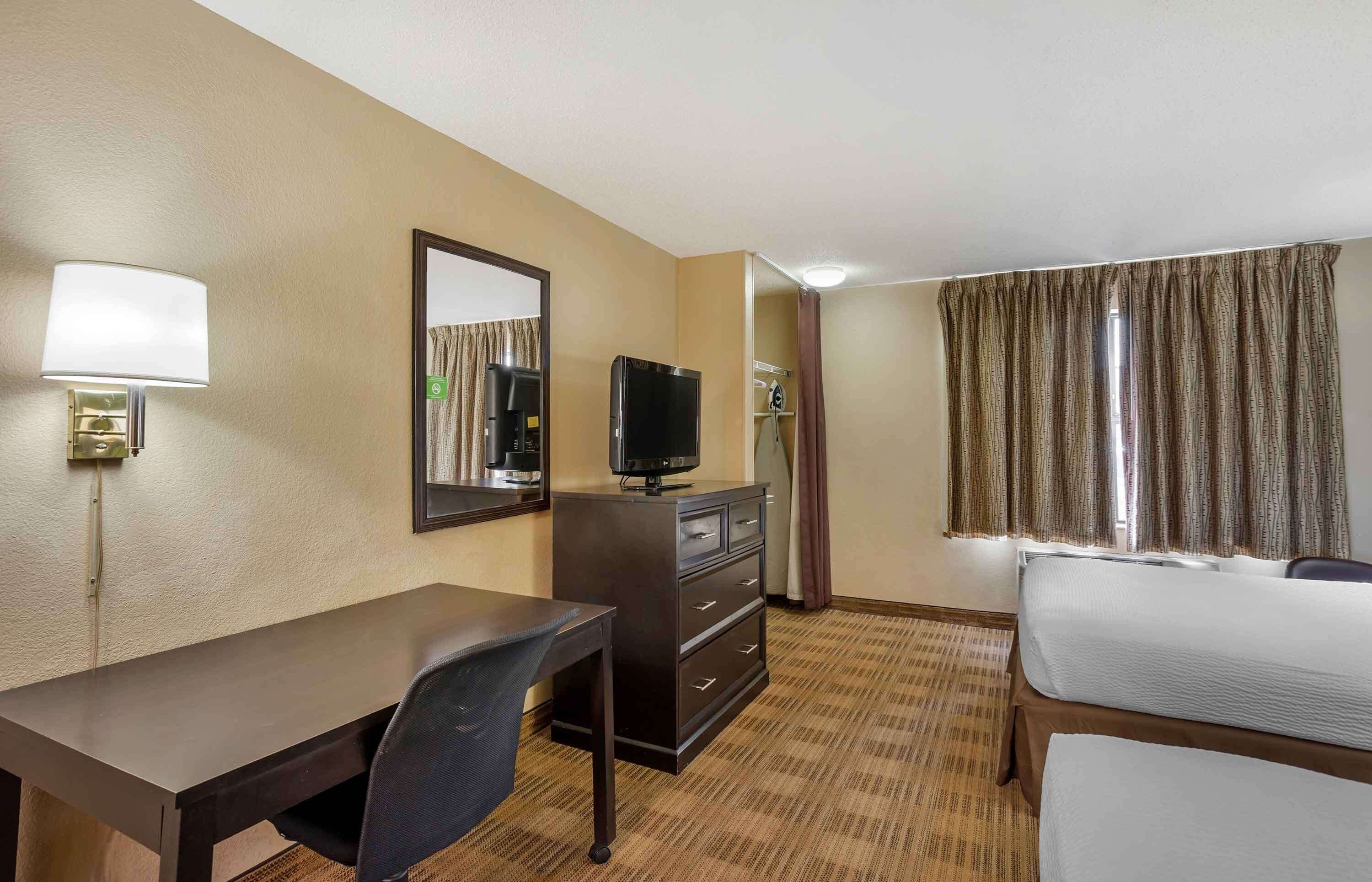 Quarto de hotel com escrivaninha, cadeira, televisão, cômoda e cama.