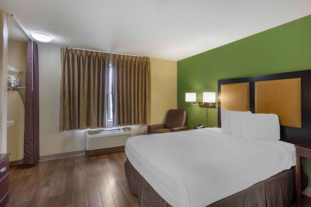 A hotel room with a large bed, a chair, a desk, and a window with curtains.
