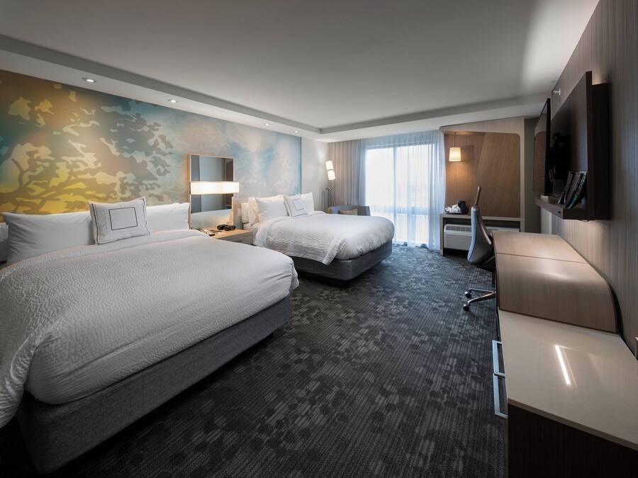A hotel room with two beds, a desk, a chair, a TV, and a large window.