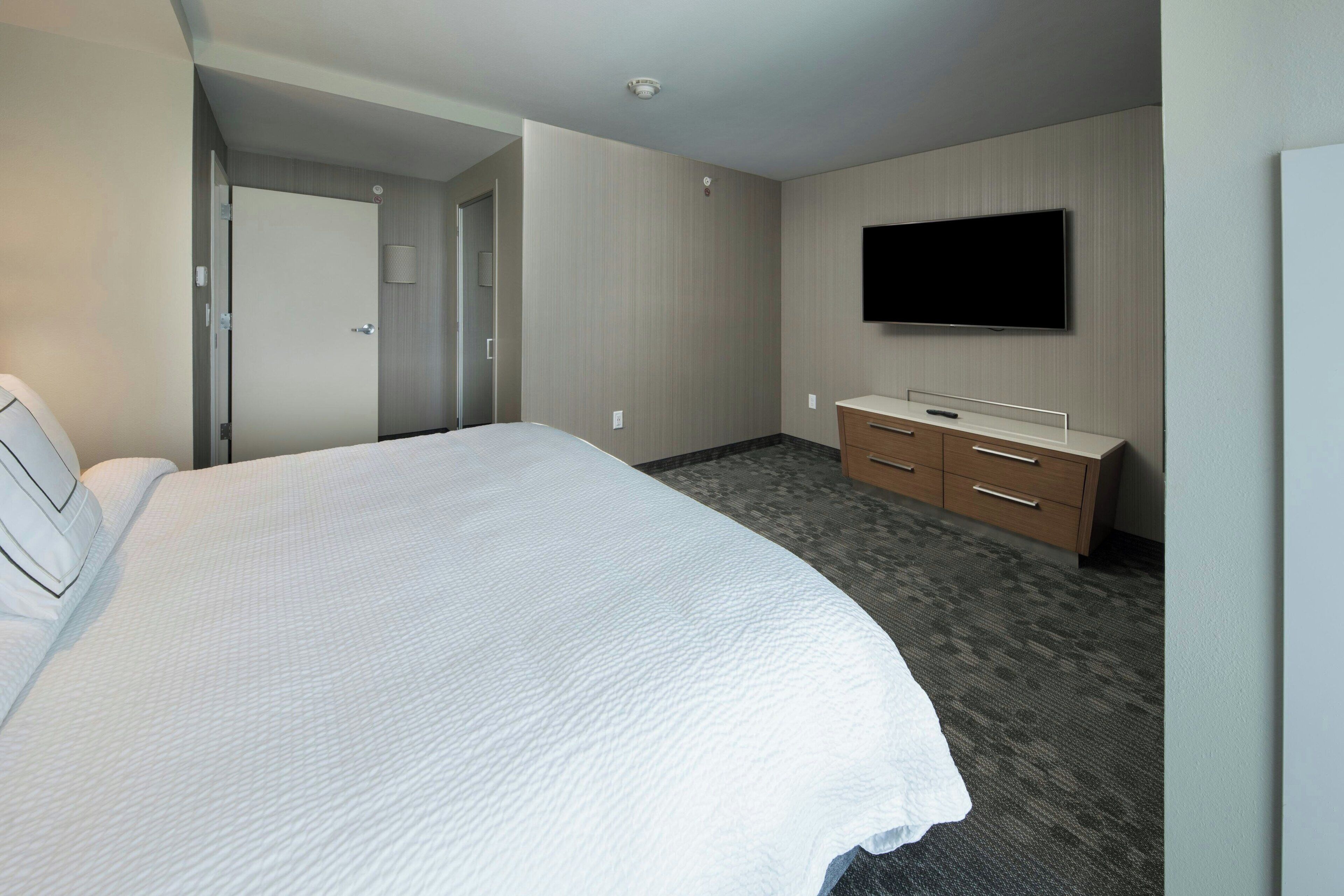 A hotel room with a large bed, a TV mounted on the wall, a wooden dresser, and a door leading to another room.