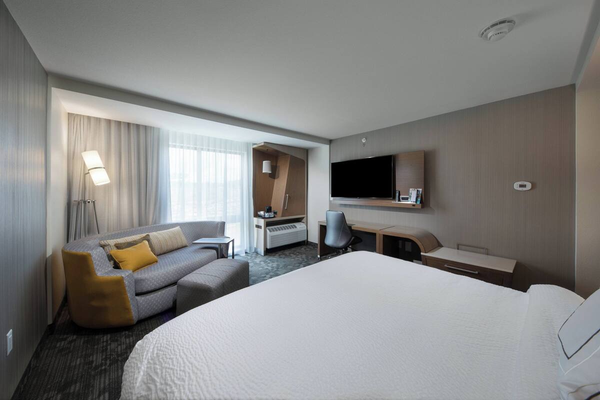 Ein Hotelzimmer mit einem großen Bett, einer Sitzecke, einem Schreibtisch, einem Sessel, einem Fernseher und einem Fenster mit Vorhängen.