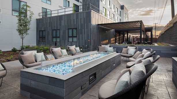 A modern outdoor lounge area with a rectangular fire feature, comfortable seating, and a building with large windows and a balcony. - Courtyard by Marriott Russellville (Russellville)