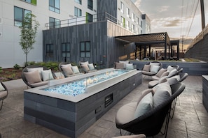 Ein moderner Außenbereich mit Lounge-Möbeln, einem rechteckigen Feuerornament und einem Gebäude mit großen Fenstern und einem Balkon.