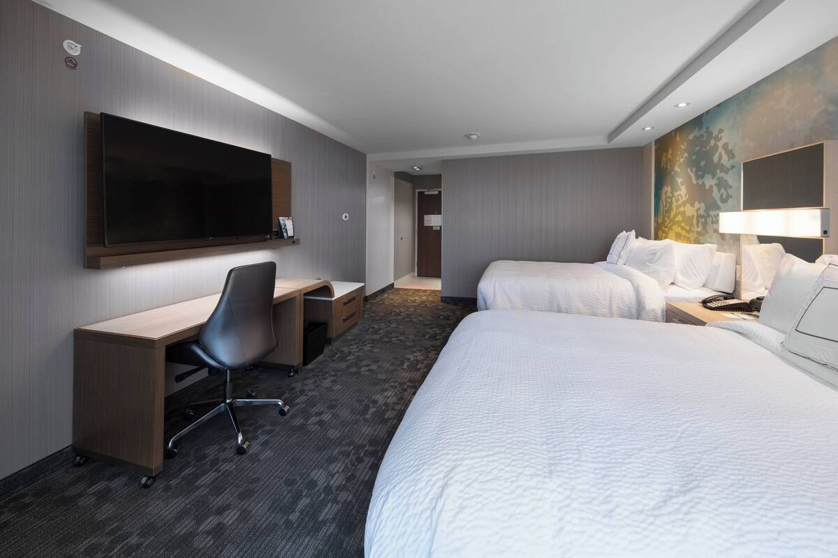 Ein Hotelzimmer mit großem Flachbildfernseher, einem Schreibtisch mit Stuhl, zwei Betten und einem Nachttisch mit Lampe.