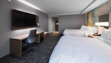A hotel room with a large flat-screen TV, a desk with a chair, two beds, and a nightstand with a lamp.