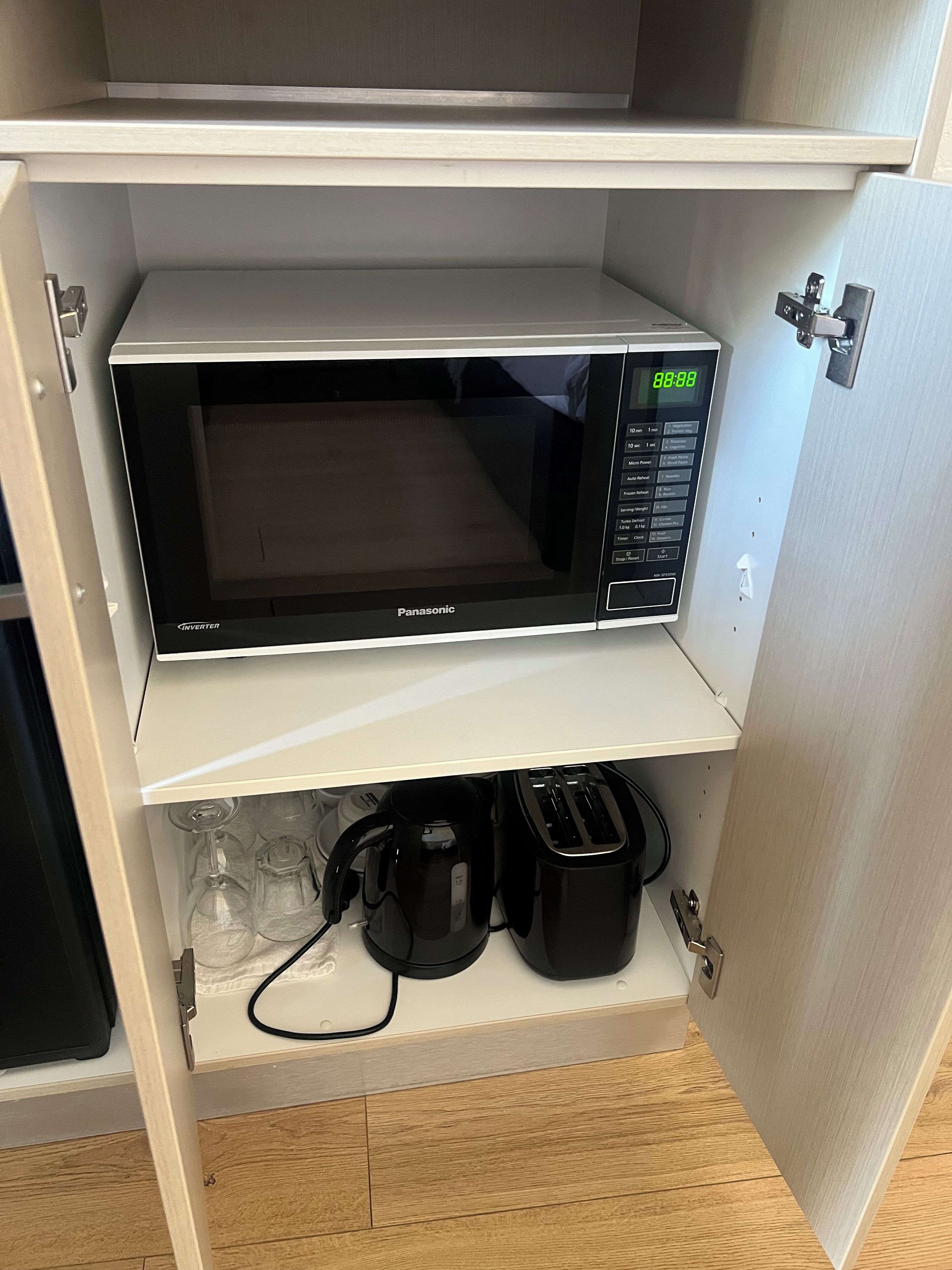 Um forno micro-ondas com visor digital e painel de controle, colocado numa prateleira acima de um armário que contém uma cafeteira, torradeira e copos de vinho.