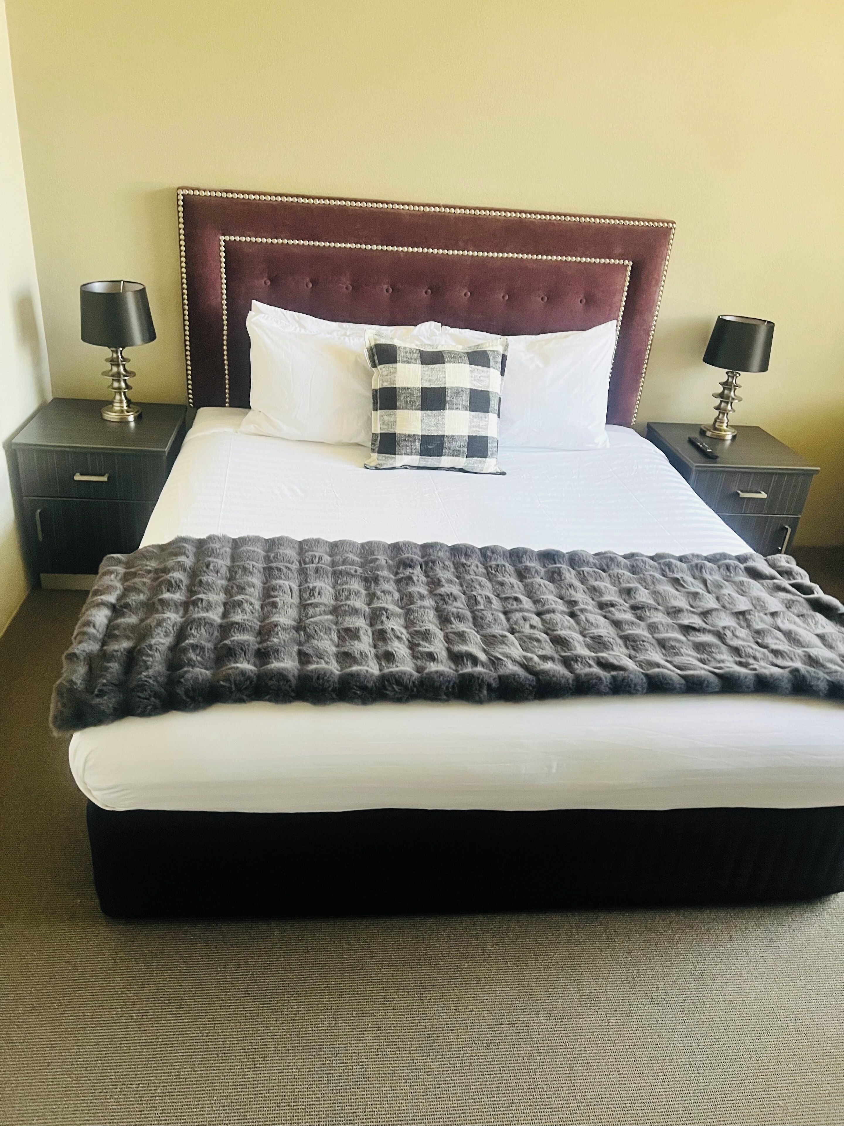 A neatly made bed with a checkered pillow, two bedside tables with lamps, and a dark headboard.