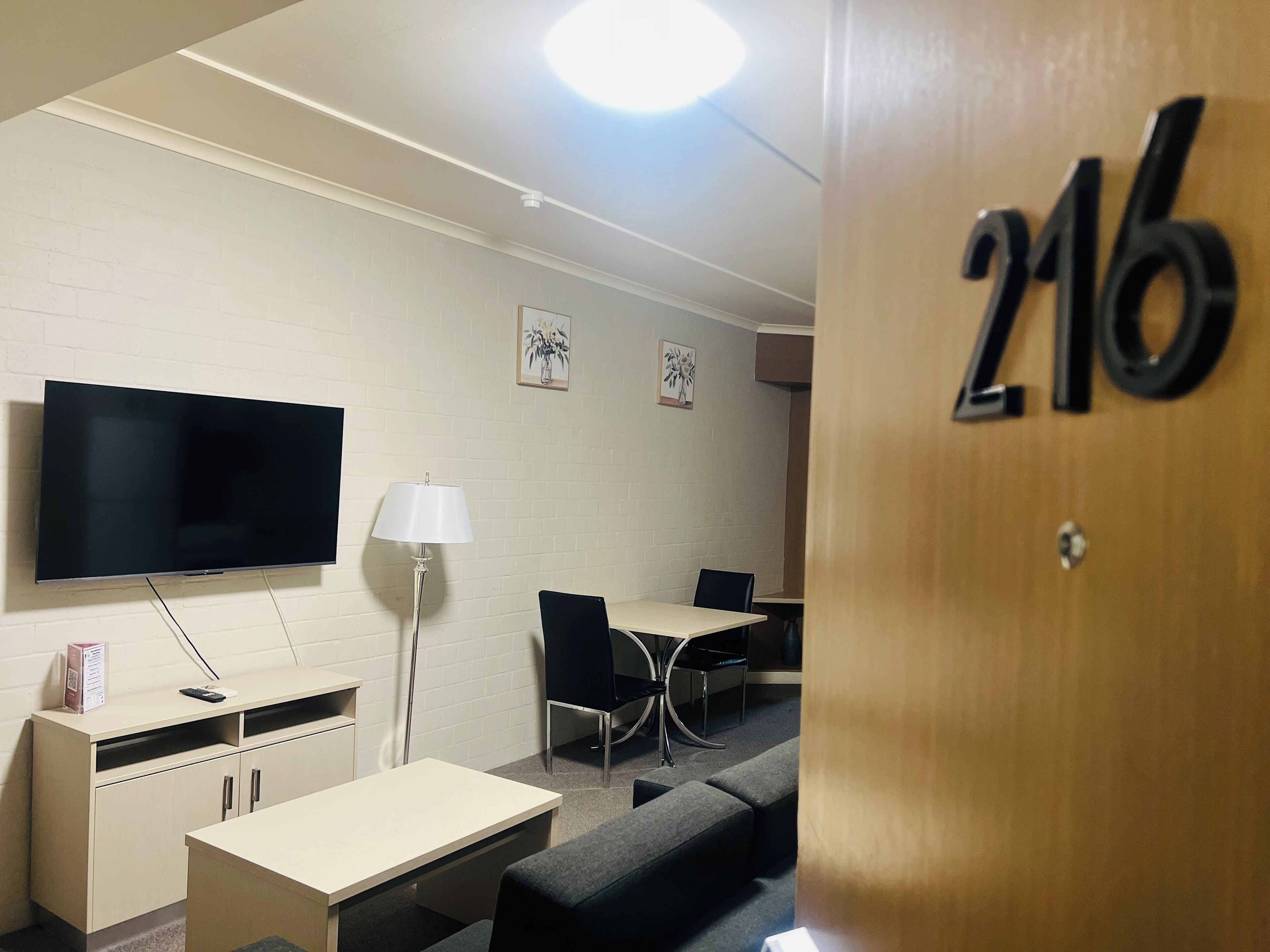 A modern room with a flat-screen TV, a desk, a chair, a sofa, and a lamp. The room has a white brick wall and a door with the number 216.