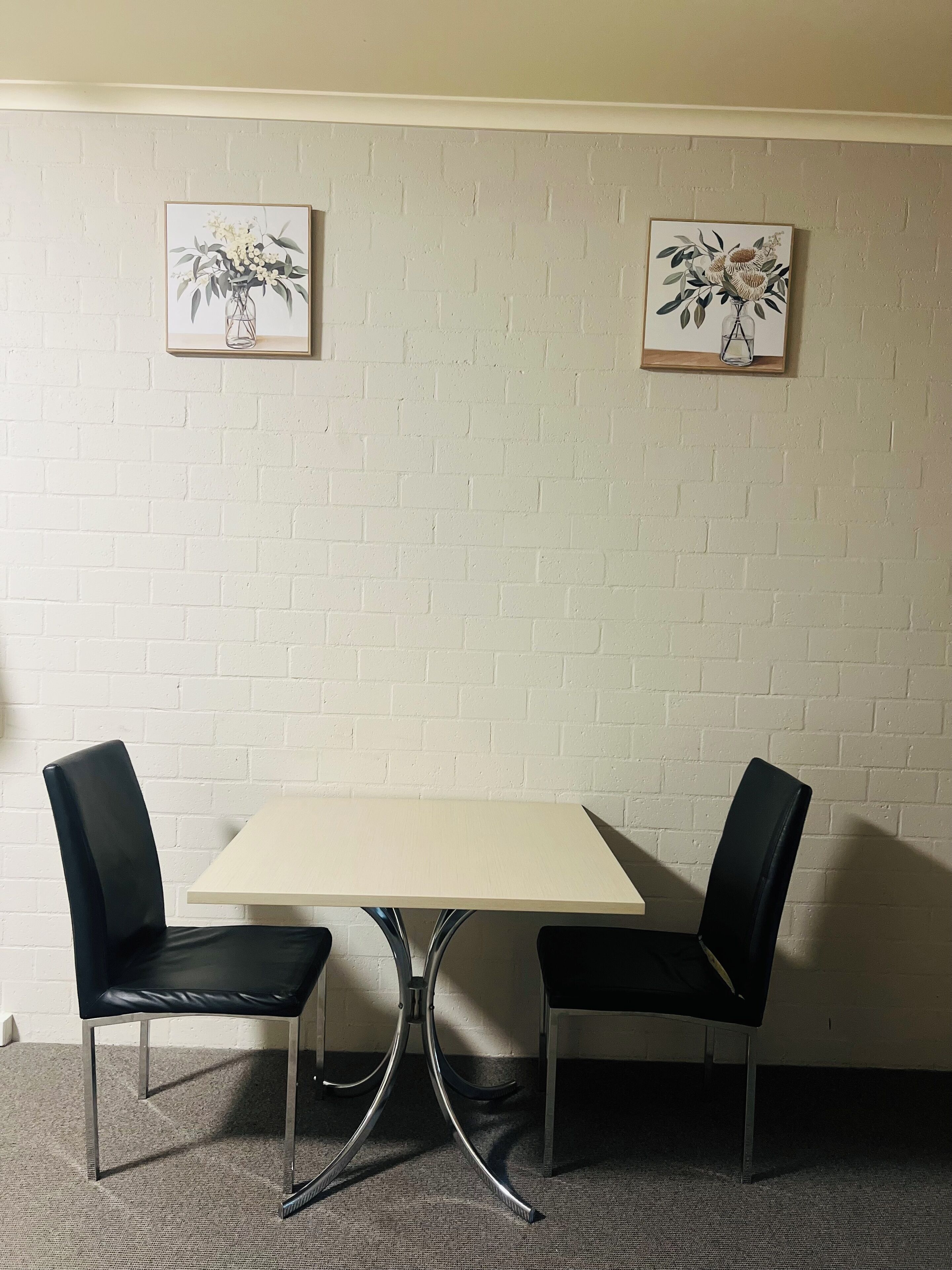 Une pièce avec un mur en briques blanches, deux chaises noires, une table de couleur claire et deux tableaux représentant des motifs floraux, encadrés.