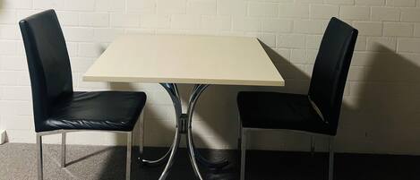 Une pièce avec un mur en briques blanches, deux chaises noires, une table de couleur claire et deux tableaux représentant des motifs floraux, encadrés.