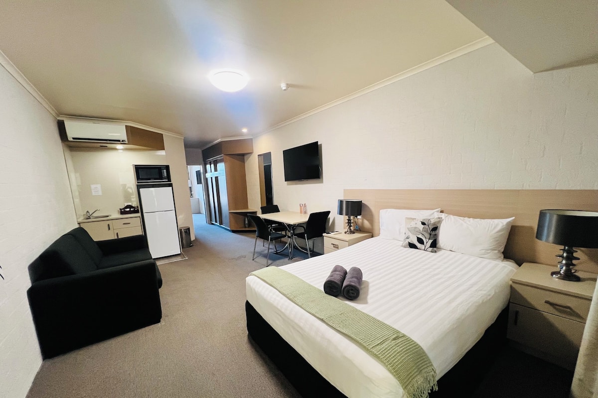 Ein Hotelzimmer mit Bett, Nachttisch, Lampe und einem Schreibtisch mit Stuhl. In der Ecke befindet sich eine Küchenzeile und ein an der Wand befestigtes Fernsehgerät.