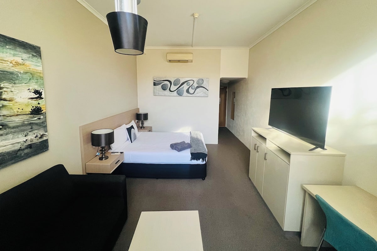 Ein Hotelzimmer mit einem Bett, Nachttisch, Lampe, Fernseher und Kunstwerken an den Wänden.