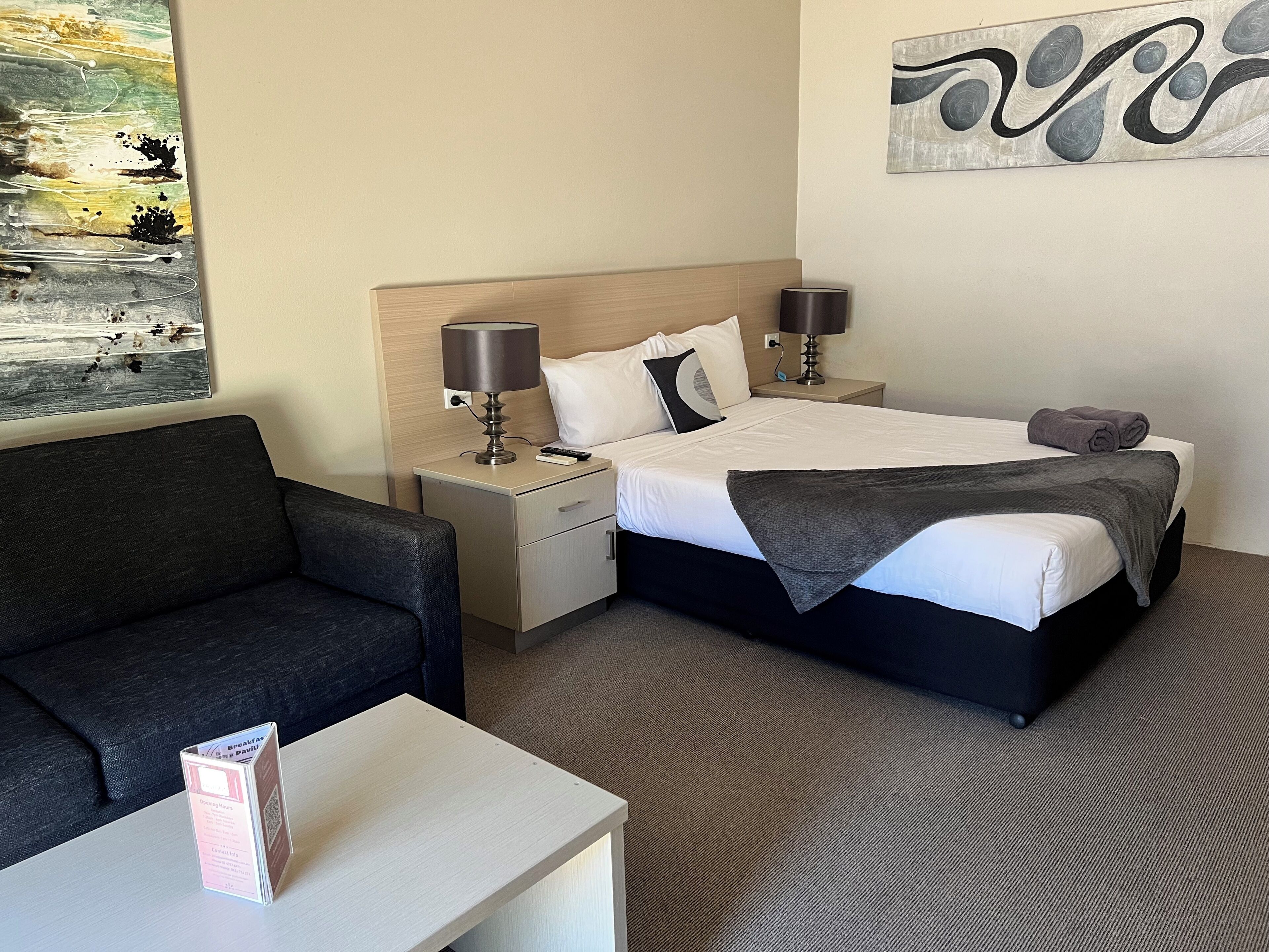 A hotel room with a bed, two bedside tables, a lamp, a remote control, a folded towel, a dark sofa, and a small table with a menu.