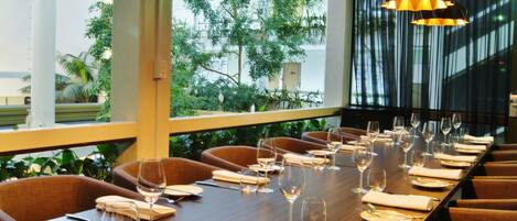 A dining area with a long table set for a meal, surrounded by chairs, with a view of greenery outside.