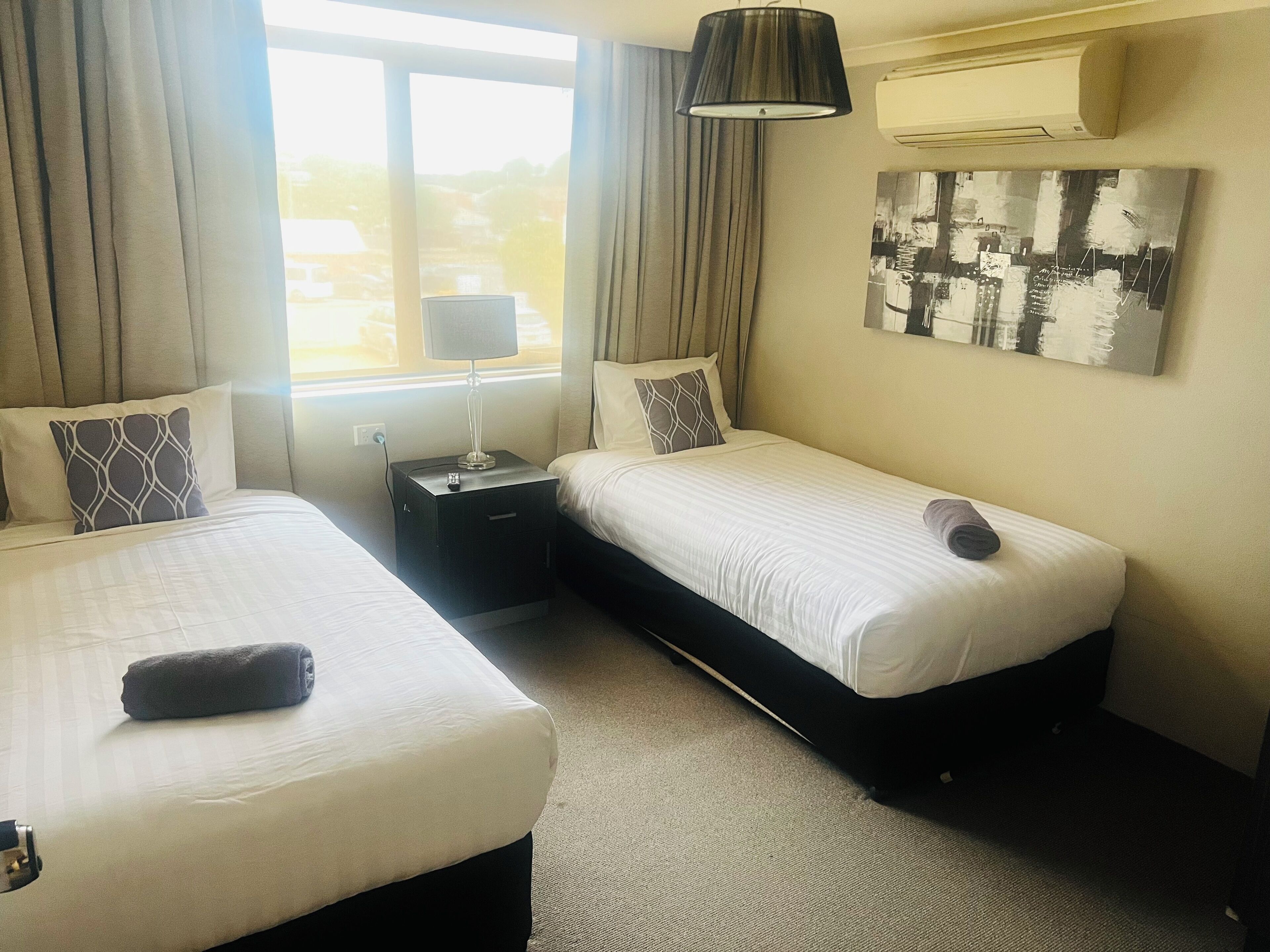 Habitación de hotel con dos camas, una mesita de noche, una lámpara, aire acondicionado y un cuadro en la pared.
