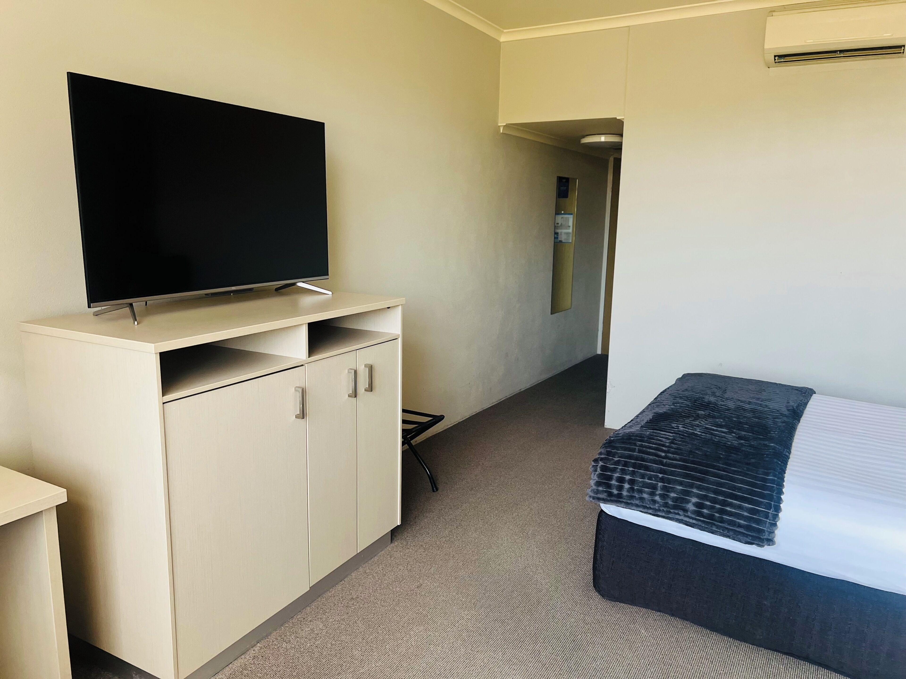 A hotel room with a flat-screen TV on a light-colored cabinet, a bed with a dark headboard, and a carpeted floor.