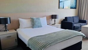 A hotel room with a bed, two bedside tables, a chair, a small table, and a painting on the wall. - Pavilion Hotel Wagga Wagga (Wagga Wagga)
