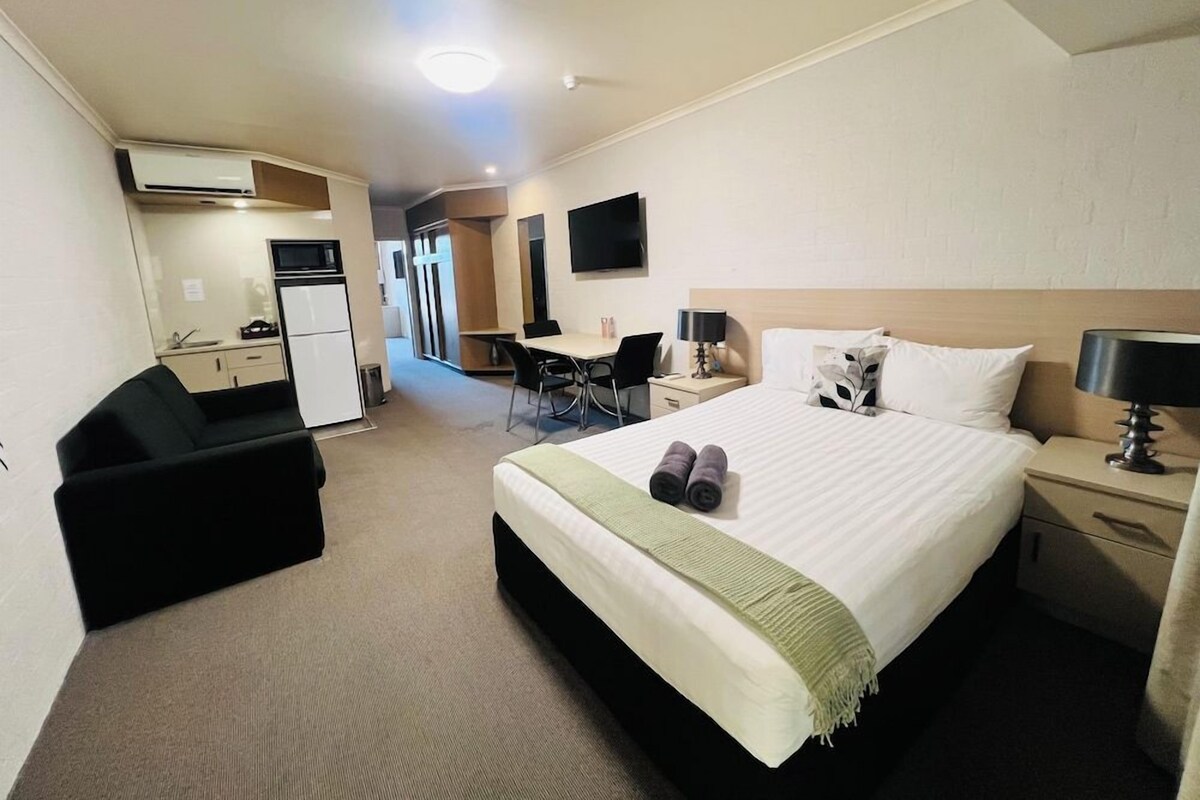 Habitación de hotel con cama, escritorio, silla, kitchenette pequeña y televisor montado en la pared.