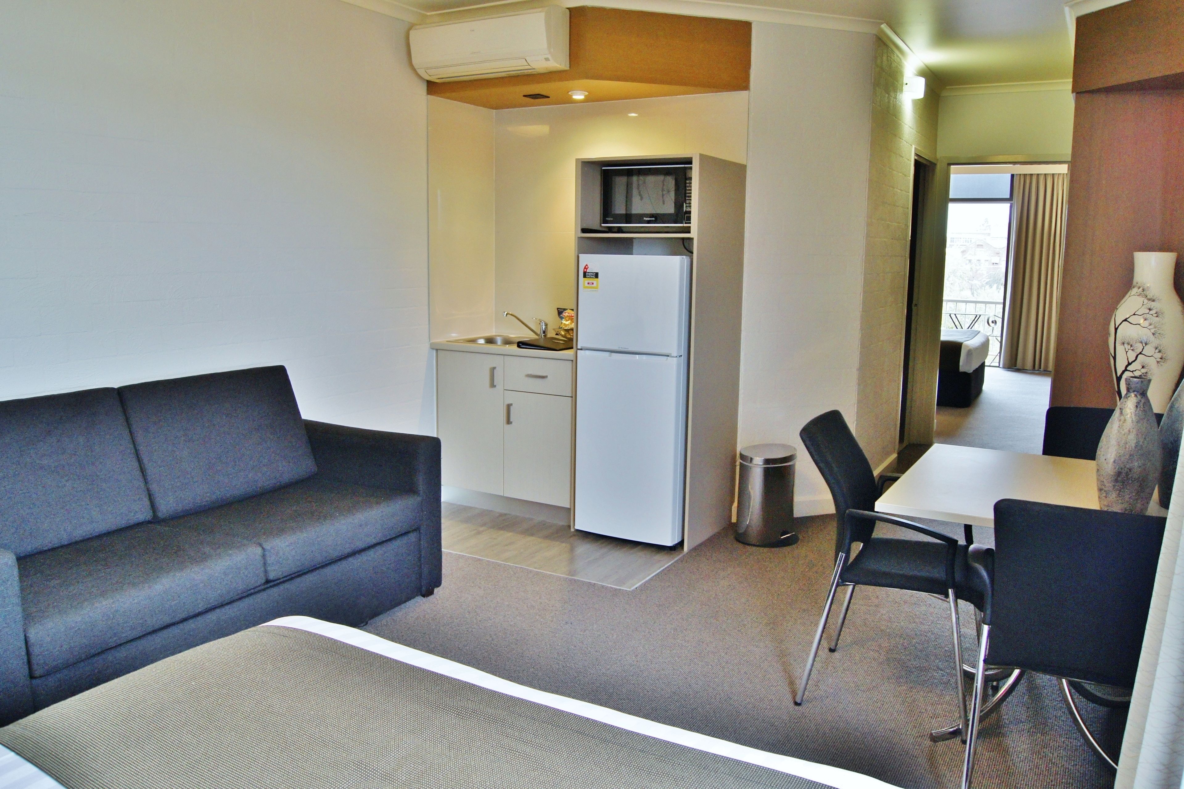 A compact hotel room with a sofa, a small kitchenette, a dining area, and a view through a window.