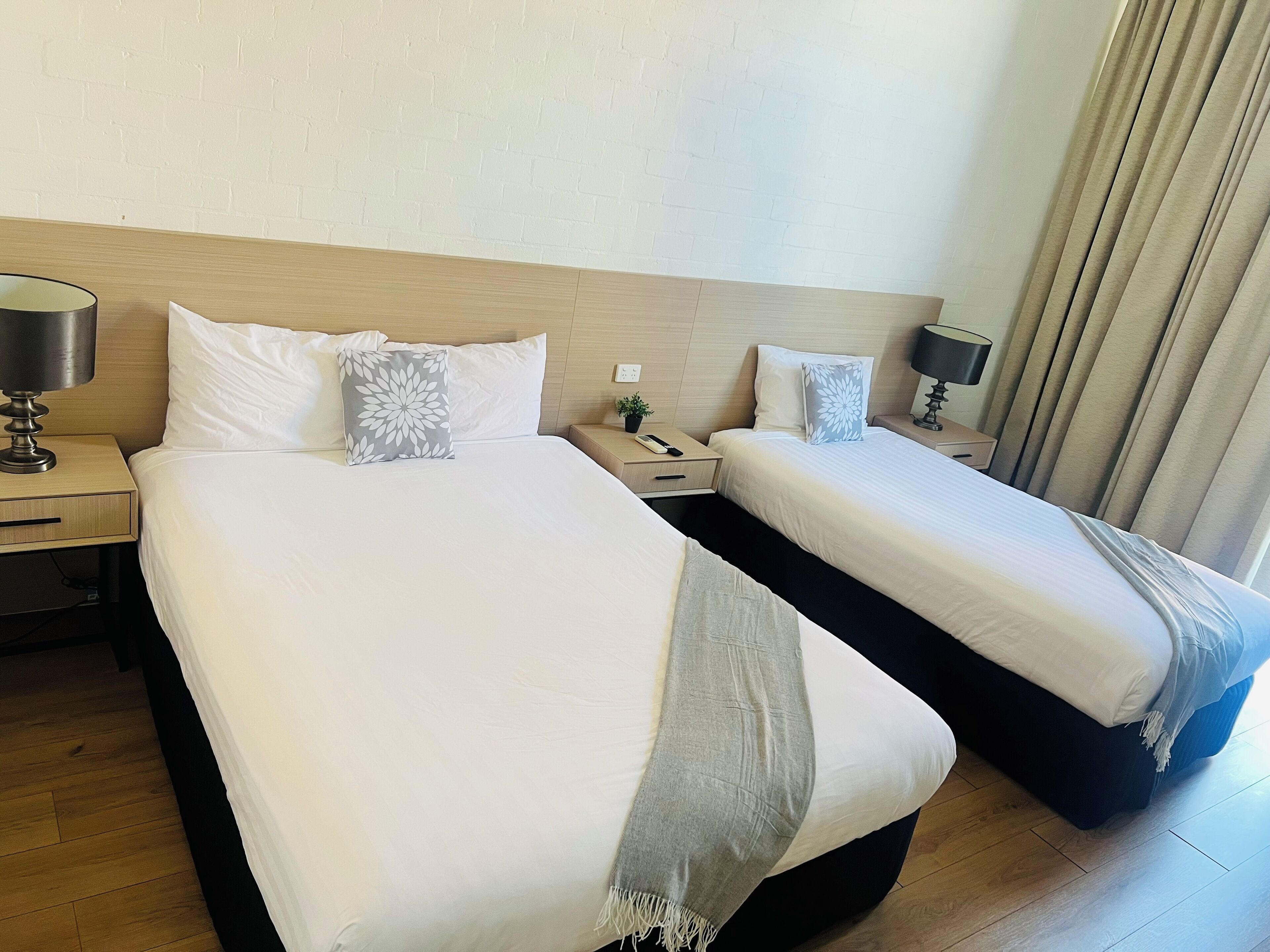 A hotel room with two beds, a nightstand with a lamp, a small table with a plant, and a window with curtains.