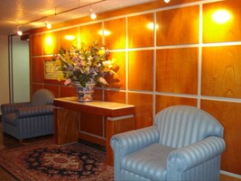 Un área de recepción con un mostrador de madera, dos sillones azules, una alfombra estampada y un jarrón con flores.