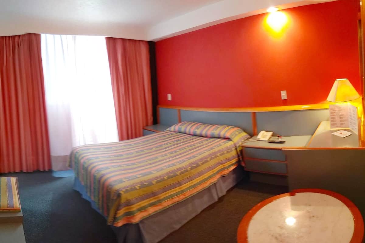 Ein Hotelzimmer mit einem Bett, einem Nachttisch mit Lampe, einem kleinen Hocker und einem Fenster mit Vorhängen.