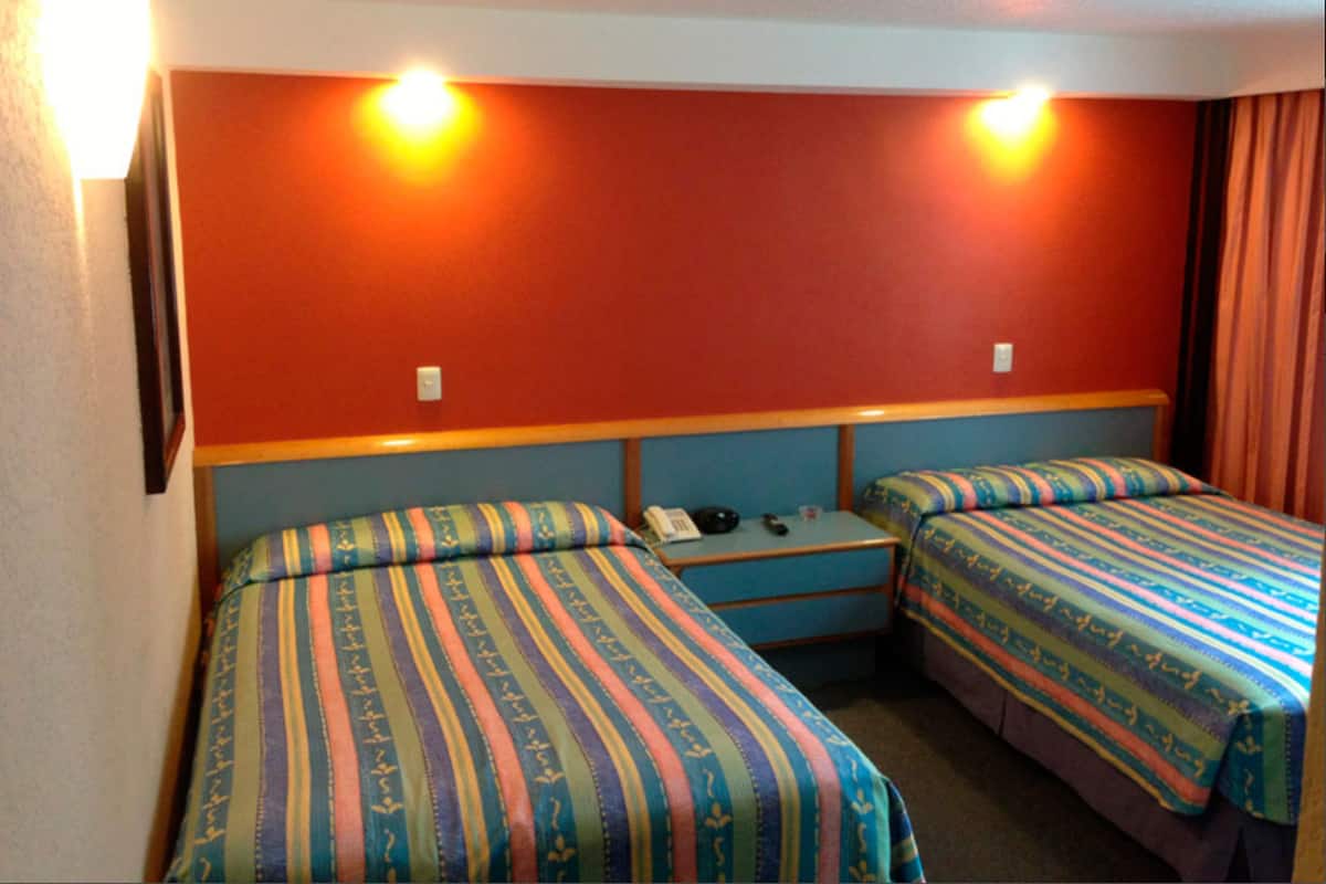 Ein Zimmer mit zwei Betten, jeweils mit gestreifter Bettwäsche, einem kleinen Tisch mit Telefon und Fernbedienung sowie einer Wandleuchte.