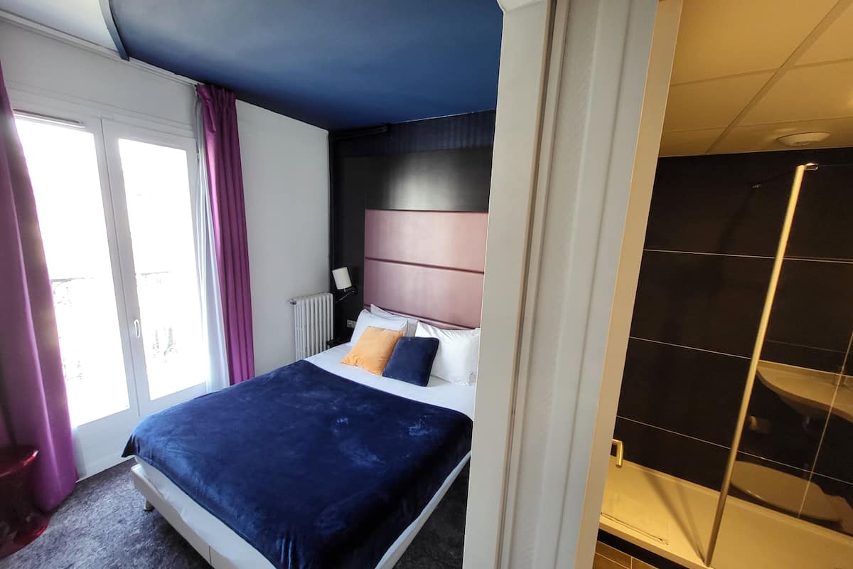 Une chambre à coucher comprenant un lit, une table de chevet, une fenêtre avec des rideaux, un radiateur et une salle de bain avec une douche.