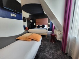 Quarto de hotel com duas camas, uma mesinha de cabeceira vermelha, televisão na parede e ventoinha no teto.