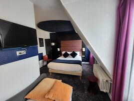 A hotel room with a bed, a TV mounted on the wall, a chair, a small table, and a radiator.