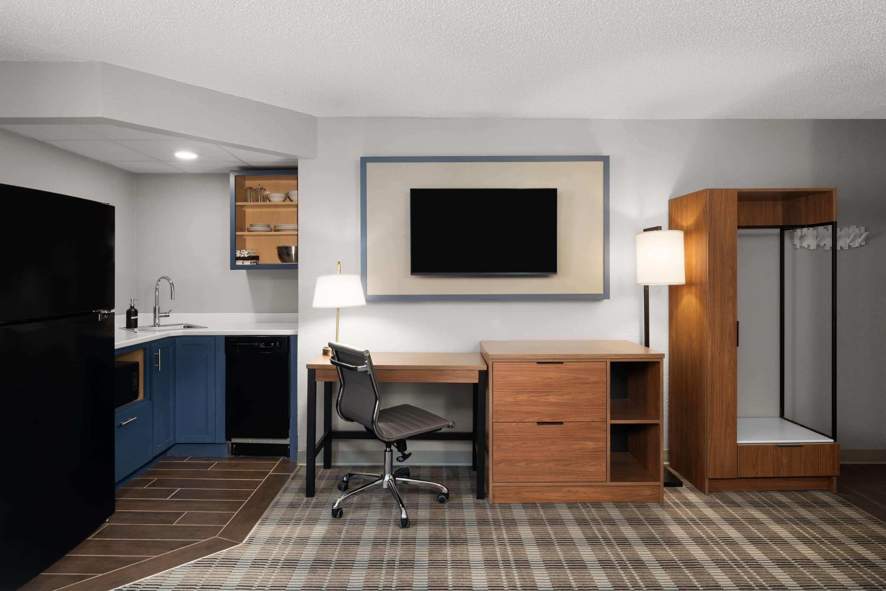 A compact hotel room with a kitchenette, a desk with a chair, a TV mounted on the wall, and a wardrobe.