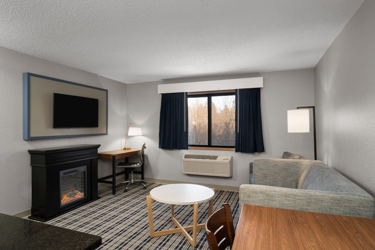 A hotel room with a fireplace, a round table, a sofa, a TV mounted on the wall, and a window with curtains.