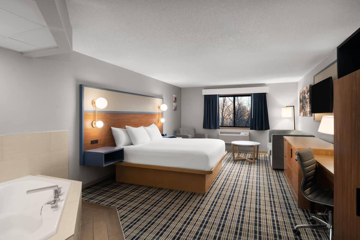 A hotel room with a large bed, a desk, a chair, a small table, a sofa, a TV, and a bathroom with a bathtub.