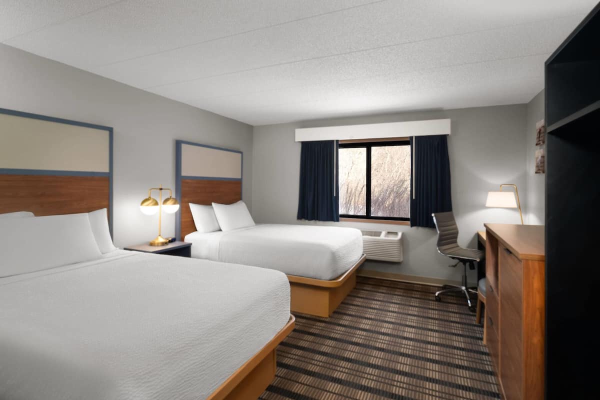 A hotel room with two beds, a desk, a chair, a lamp, and a window with curtains.