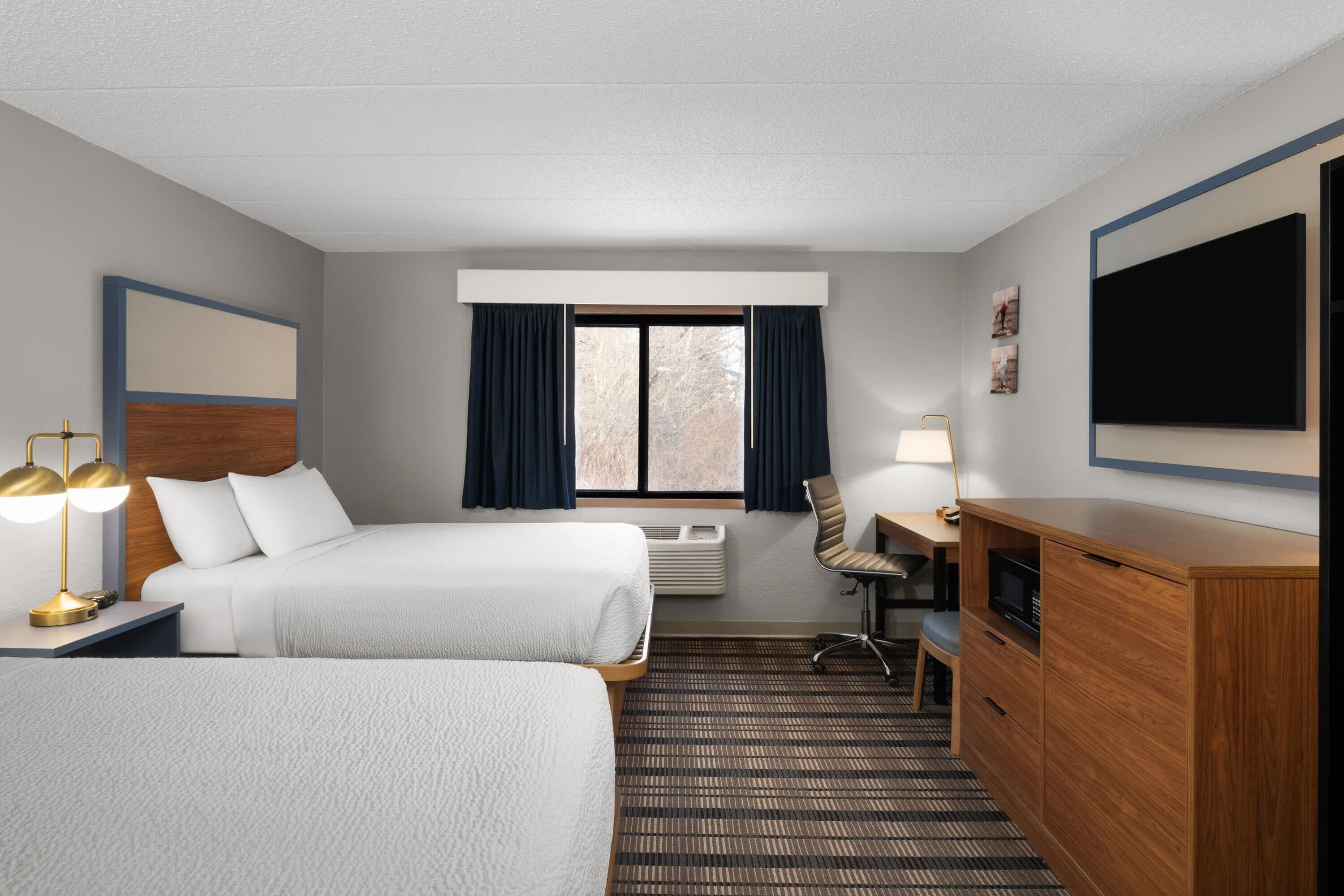 A hotel room with two beds, a desk, a chair, a TV, and a window with curtains.
